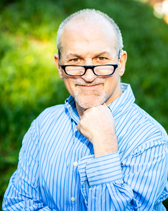 A portrait of Ability Together’s Founder, Keith Bonchek, wearing a lightly striped blue shirt, in front of a rich green background of bushes, expressing a radiating warm smile of gratitude.