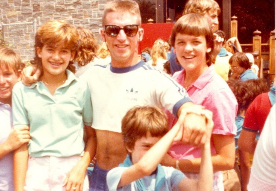 Keith as a young adult camp counsellor stand with several happy children around him. He's wearing sunglasses and smiling; the photo is typical of the 1970s.